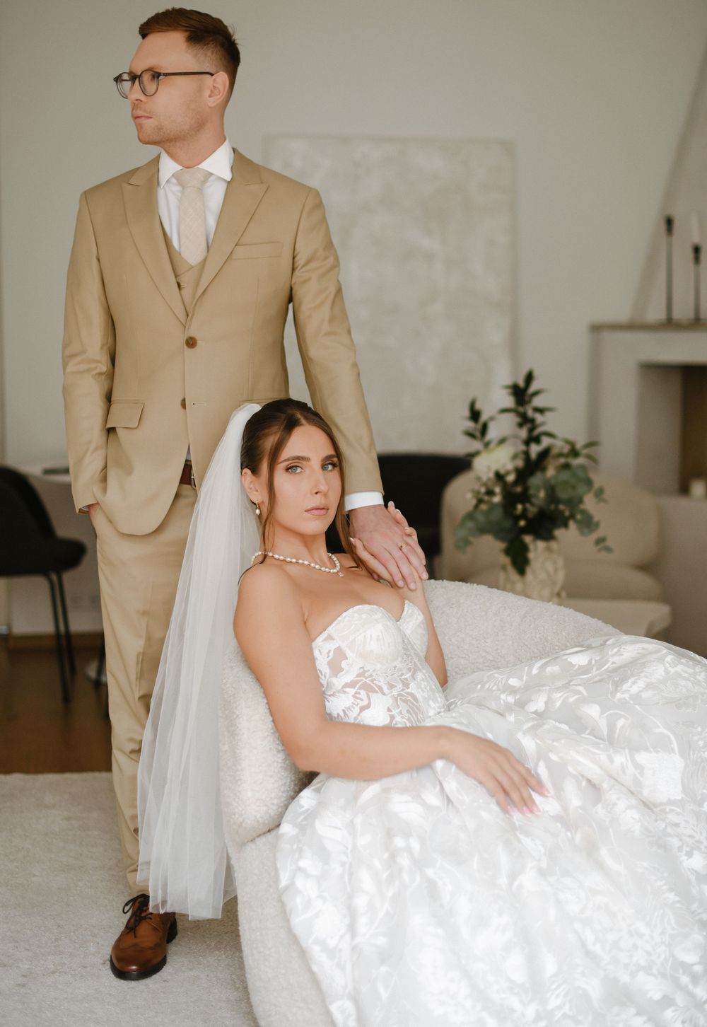 A couple in formal wedding attire poses in an elegant indoor setting with white walls and minimalist decor.