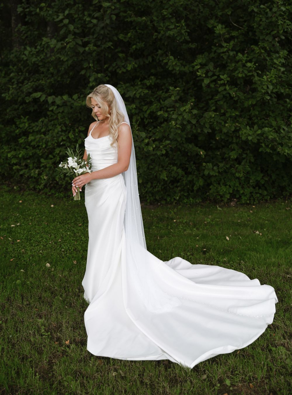 A flowing white wedding dress with long train moves elegantly across a grassy outdoor setting.