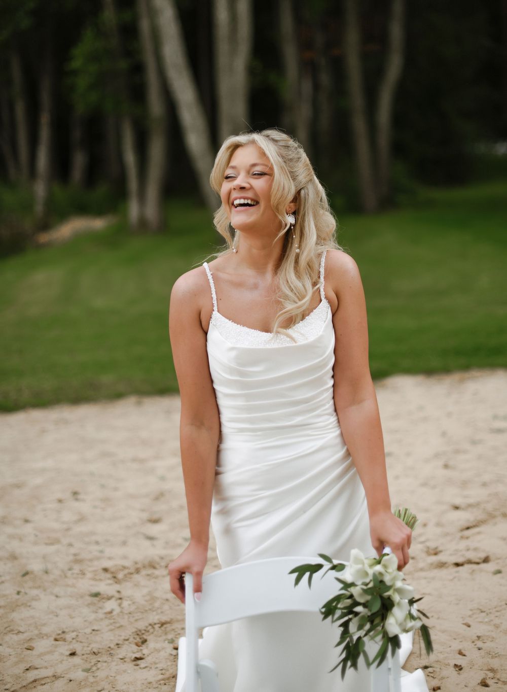 Joyful bride in white spaghetti strap wedding dress laughs while holding greenery bouquet on sandy beach path.
