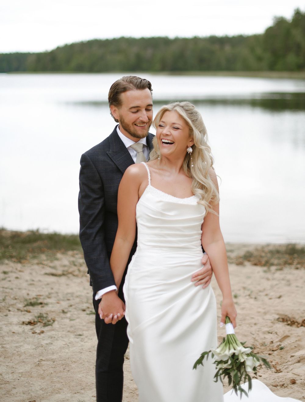 A couple shares a romantic moment on a beach near a lake, with the bride in a flowing white dress and groom in dark suit.