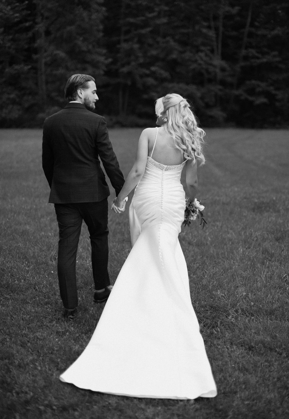 Black and white photo of a couple in wedding attire walking together on a grassy field.