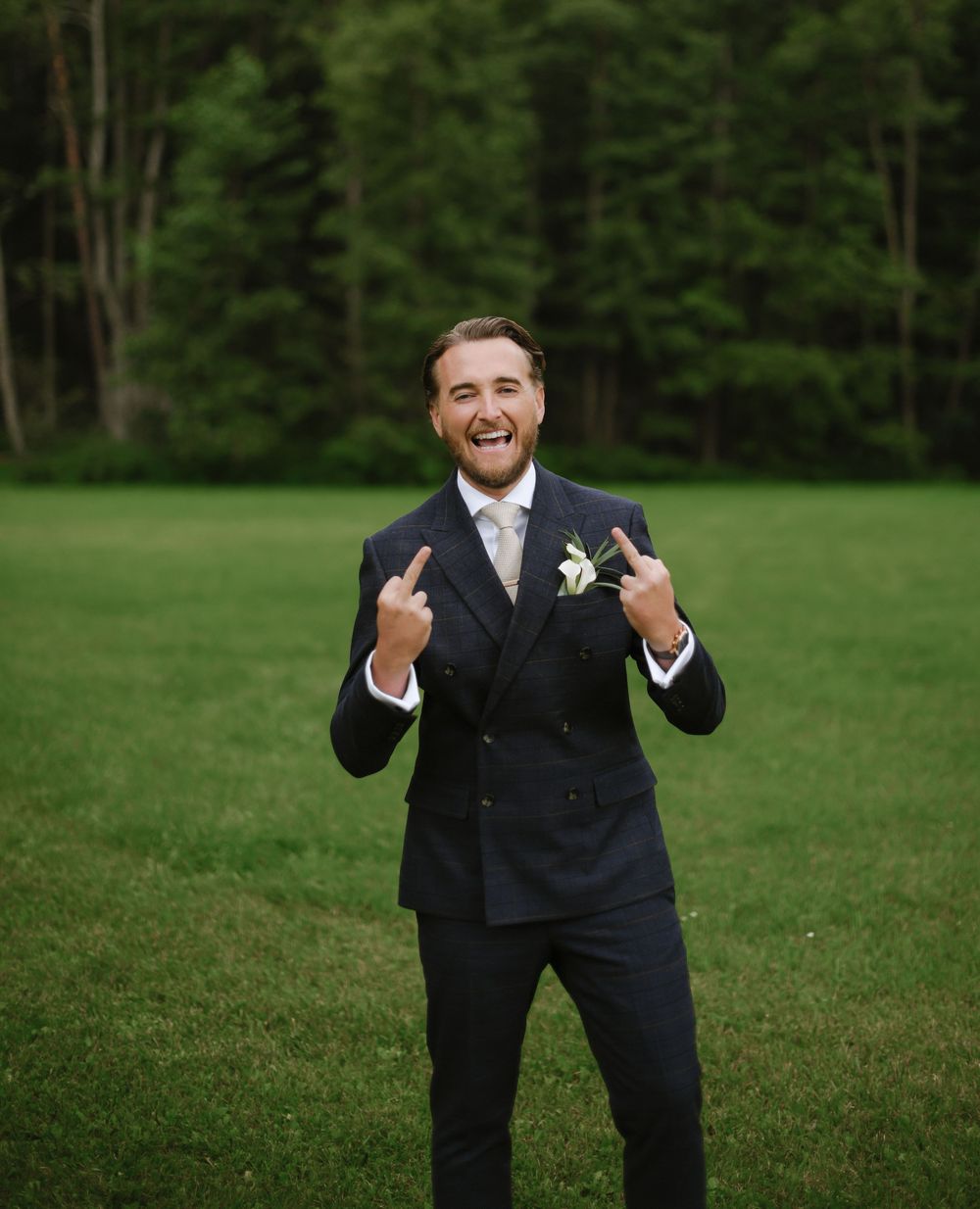 A person in a dark suit strikes playful poses in a grassy field with trees in the background.
