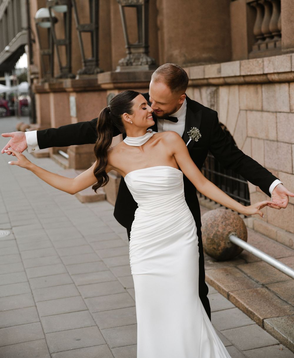 A couple in formal wedding attire share a romantic dance pose together on a stone-columned outdoor terrace.