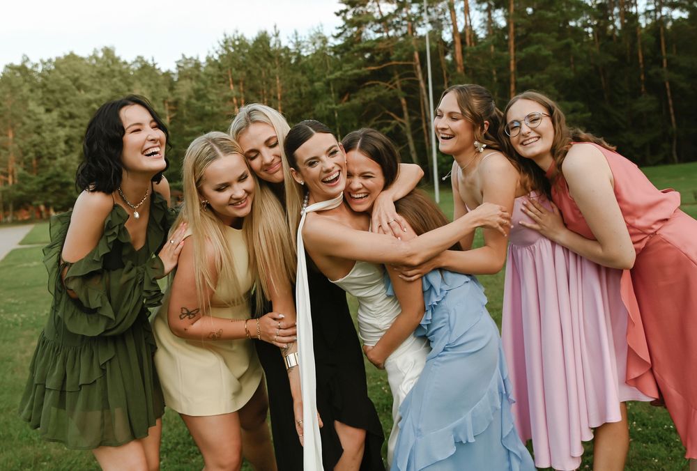 A group of friends in colorful dresses laugh together while posing outdoors with pine trees in the background.