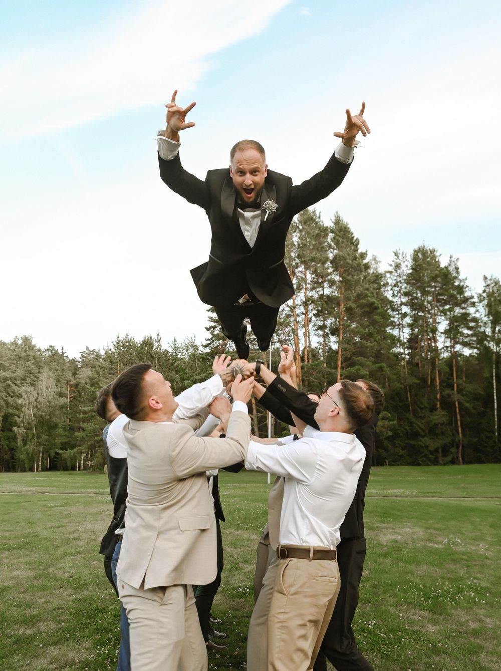 Groomsmen in formal attire playfully pose on a grassy field with trees in the background showing progression of jumping.