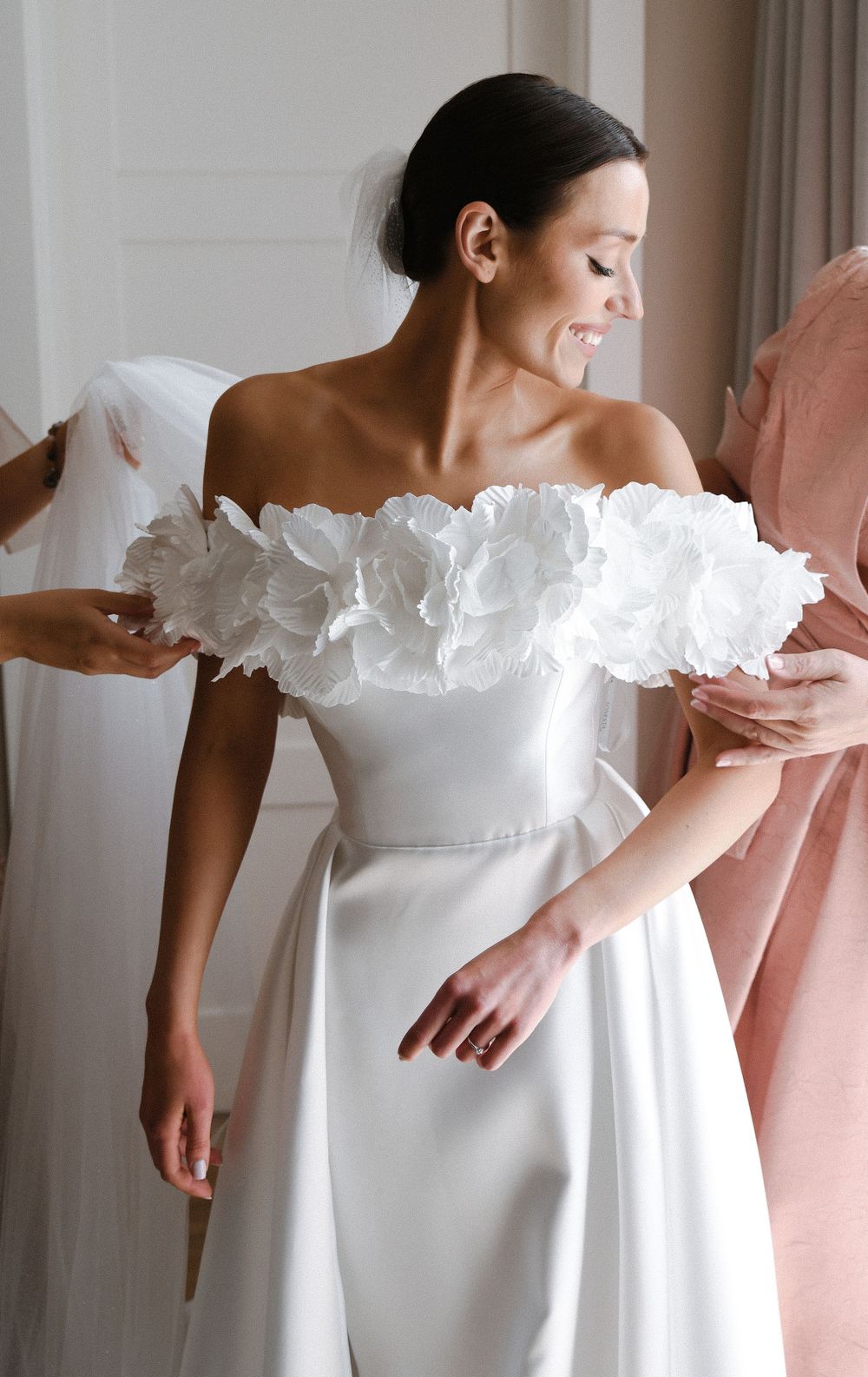 Elegant white wedding dress with dramatic floral off-shoulder neckline detail shown in bridal fitting room.