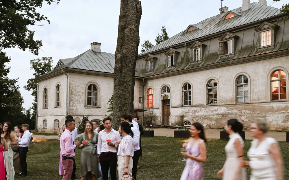 Guests mingle outside a historic mansion with arched windows and stone facade during an elegant event at sunset.
