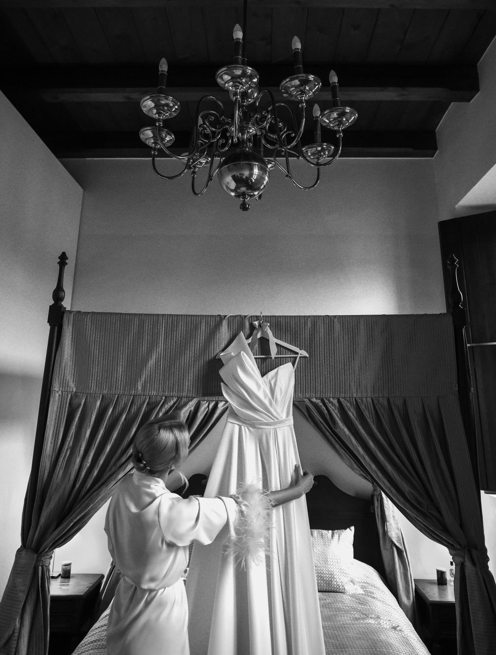 Black and white photo of wedding dress being prepared in room with chandelier.