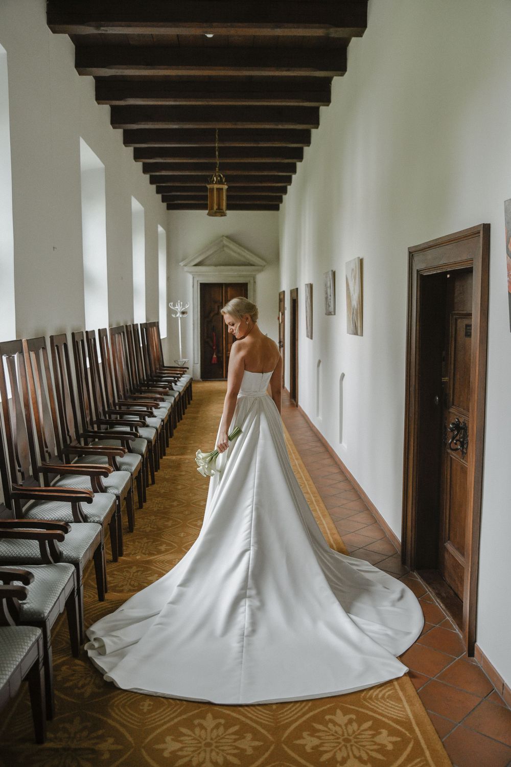 Bride in elegant white gown with long train standing in hallway with wooden chairs.