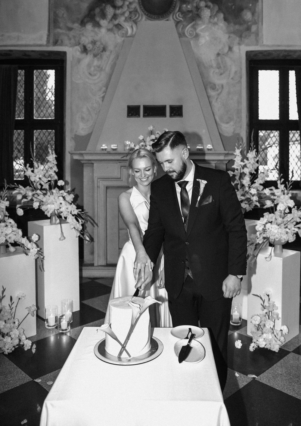 Black and white photo of couple cutting their wedding cake surrounded by floral arrangements.