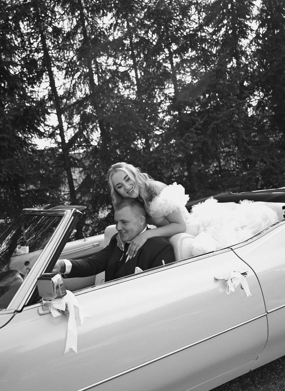 Black and white photo of a bride and groom in a classic convertible car decorated with white ribbons.