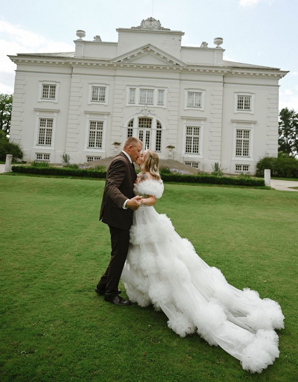 Bride and groom share a romantic kiss on lush green lawn before elegant white mansion wedding venue.