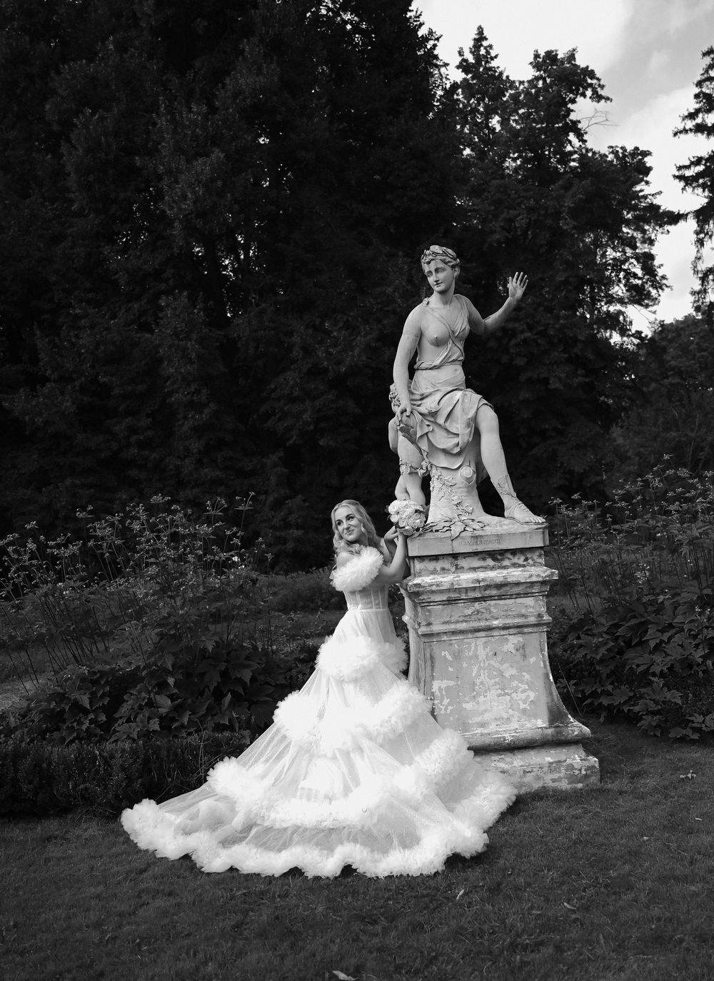 Bride in flowing white gown poses beside classical garden statue surrounded by lush trees in black and white photo.