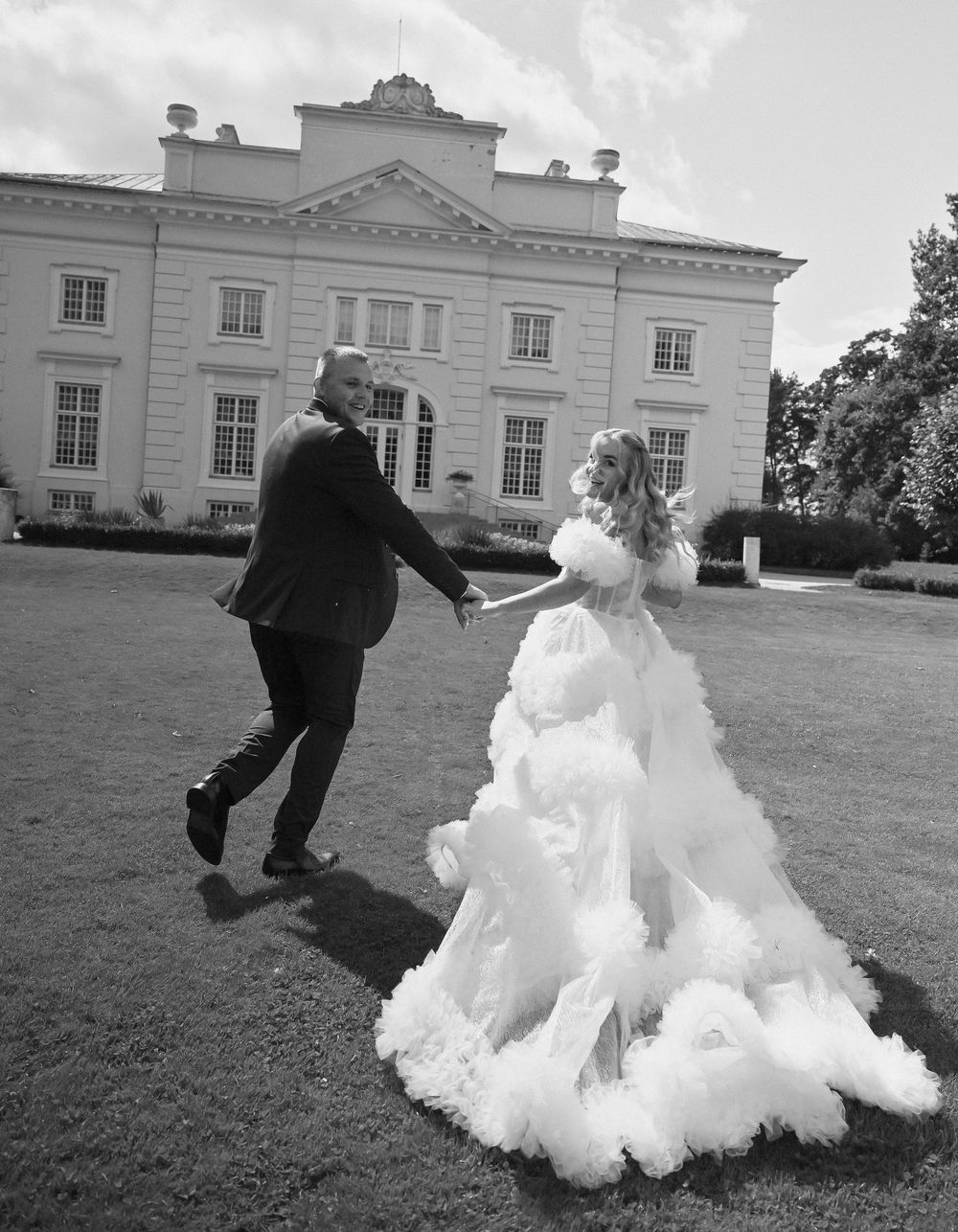 Black and white wedding photo of couple dancing on lawn before grand manor house, bride in dramatic ruffled gown.