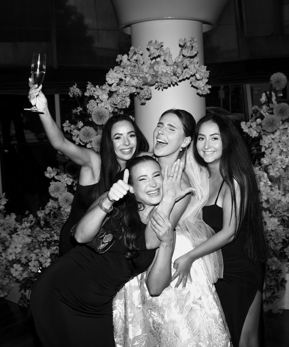 A group of partygoers celebrate joyfully with white floral decorations visible in the background.