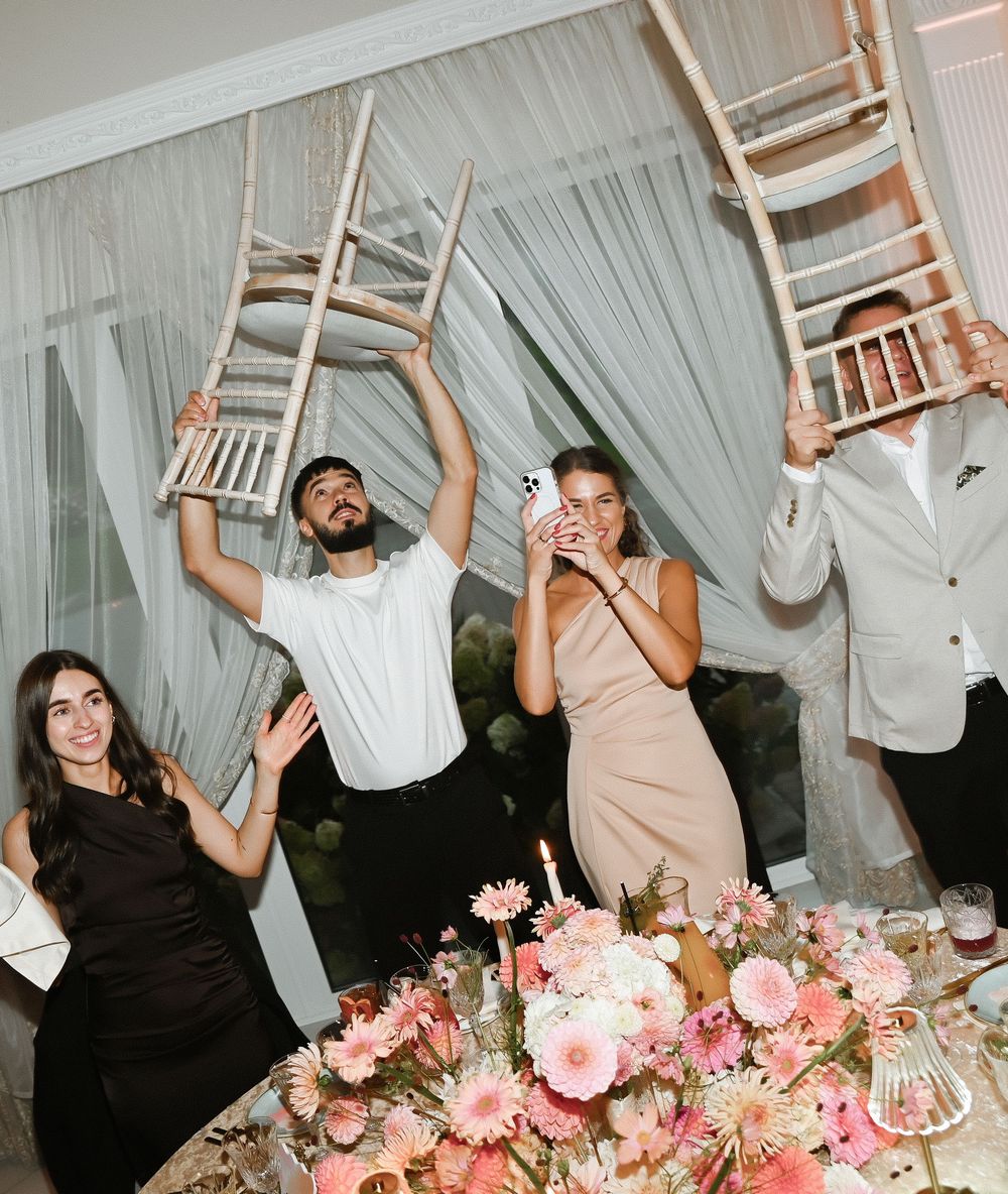 Wedding party guests celebrating with chairs raised above their heads at a reception decorated with pink floral arrangements.