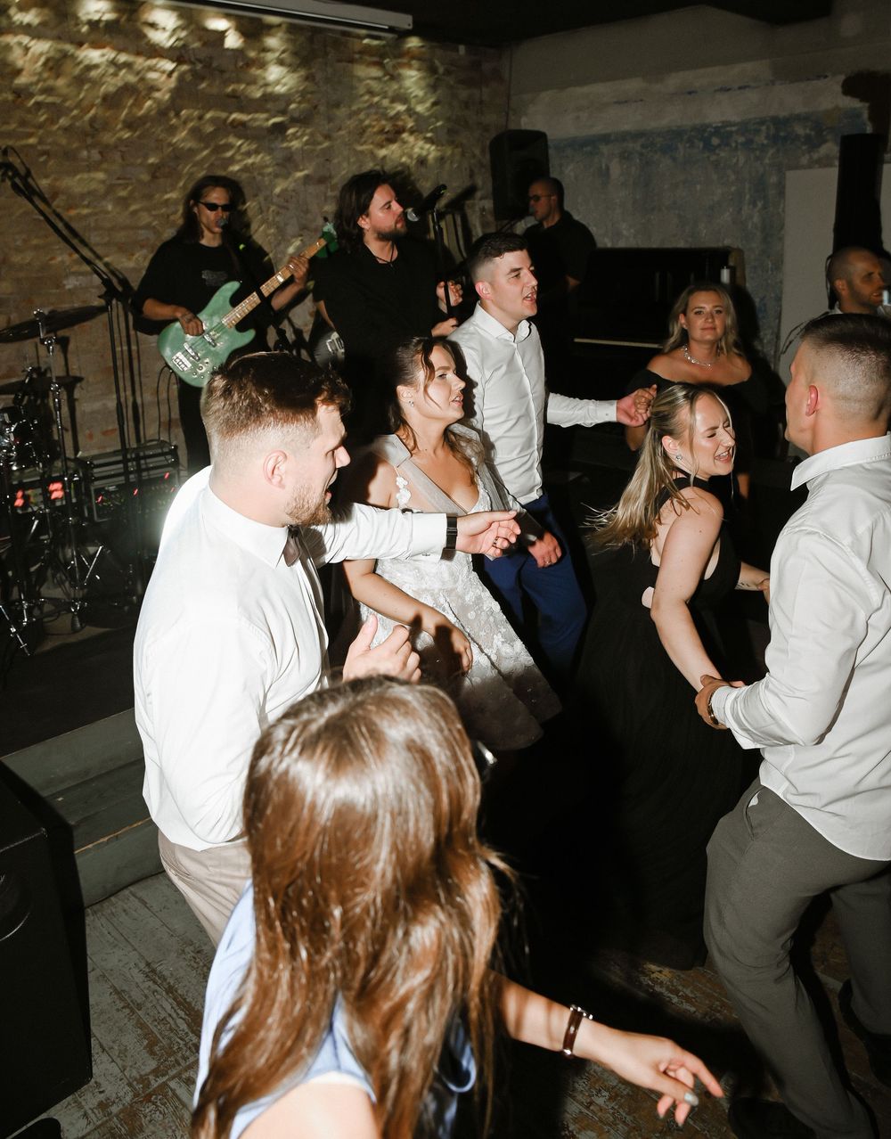 Wedding guests dancing and celebrating on the dance floor while a live band performs in the background at a reception venue.