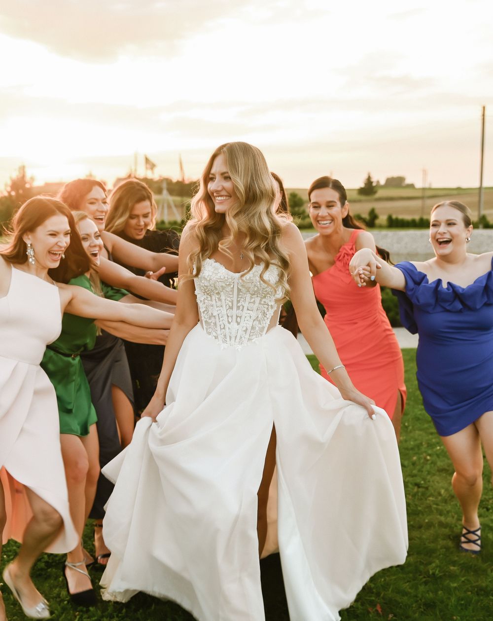 A group of bridesmaids in colorful dresses dance and twirl their white dresses in a field at sunset.