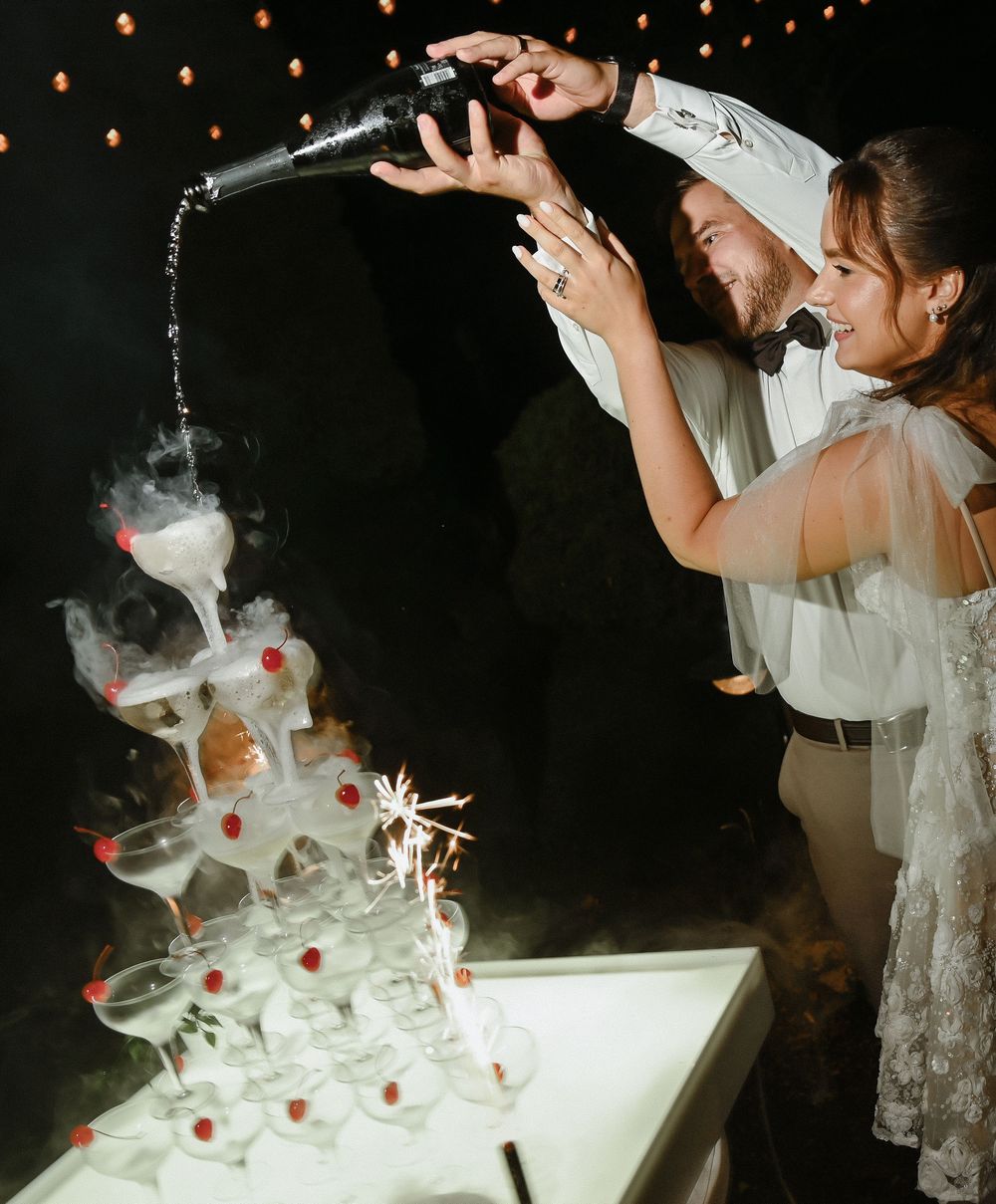 A champagne tower being poured at a wedding reception with string lights glowing in the background.