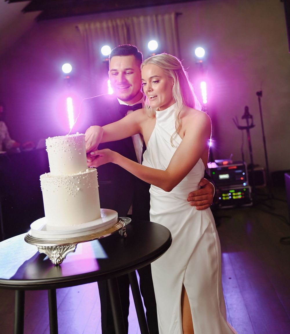 Wedding couple shares a moment near their elegant white wedding cake under purple ambient lighting at their reception.