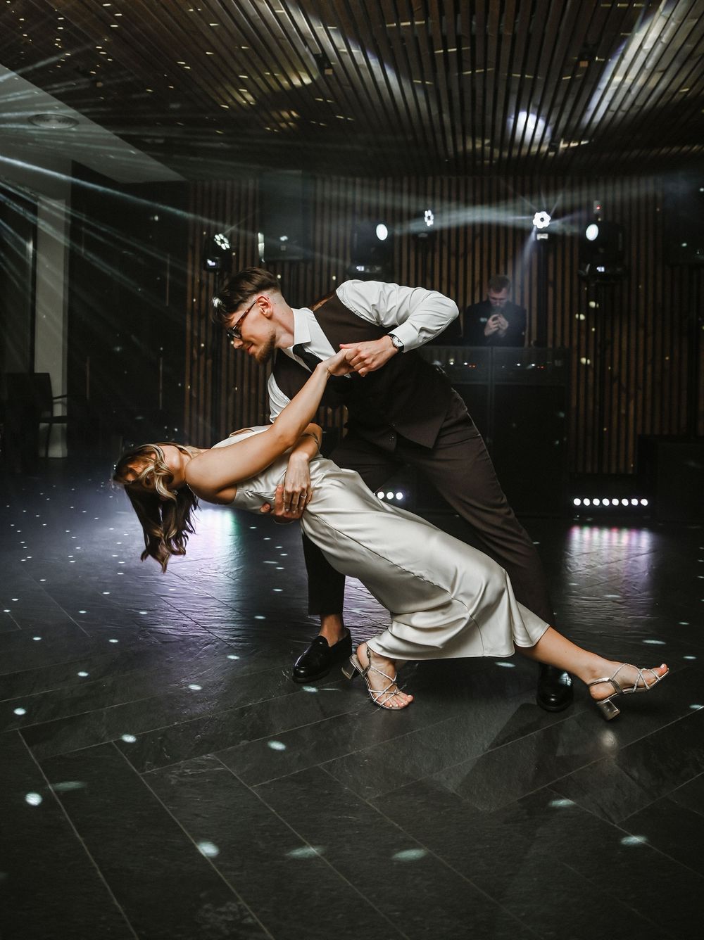 Two dancers perform a dramatic aerial dance move in a dark studio with spotlights creating a moody atmosphere.