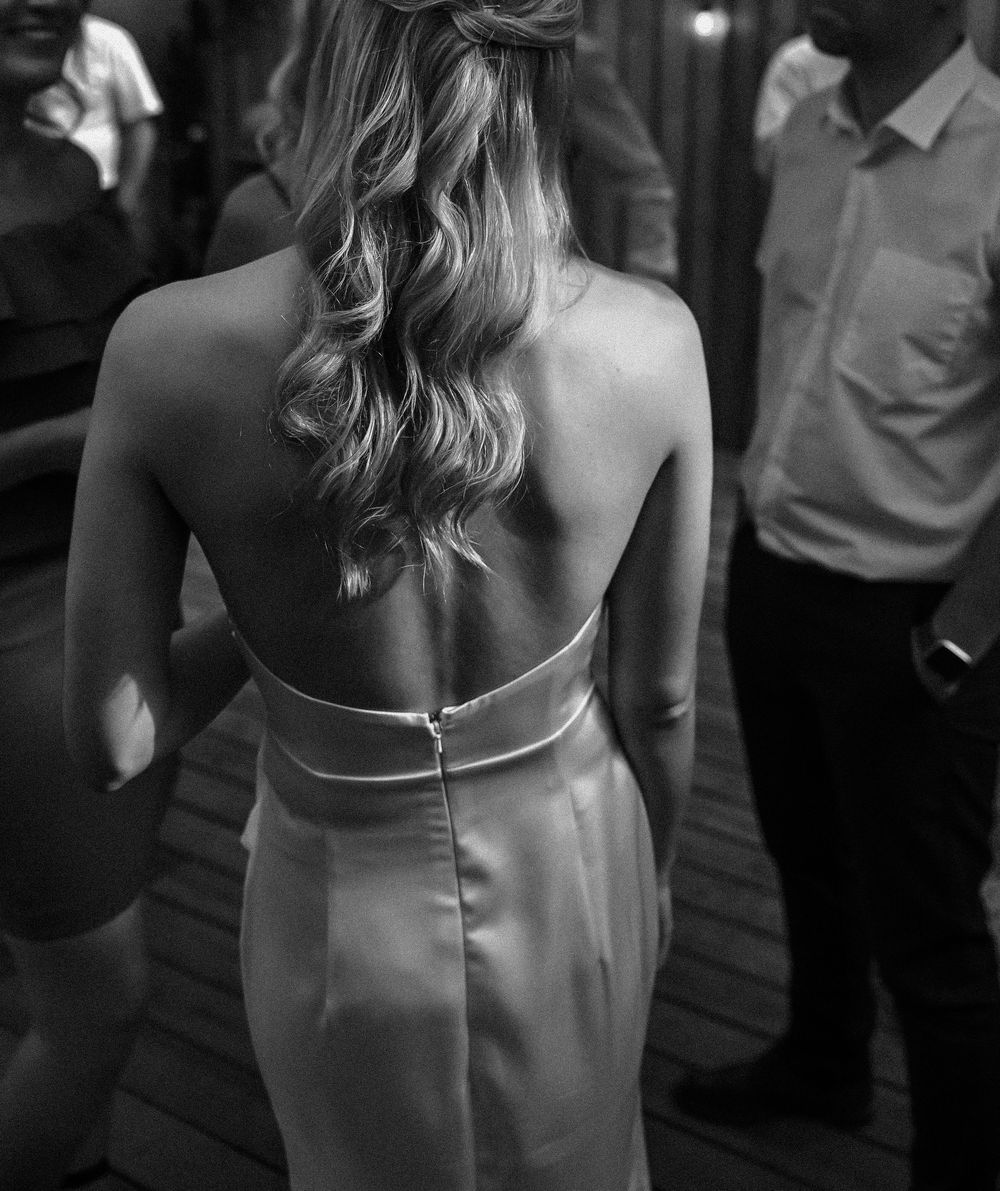 Black and white photo of a woman's back in an elegant dress with wavy blonde hair at a formal event.