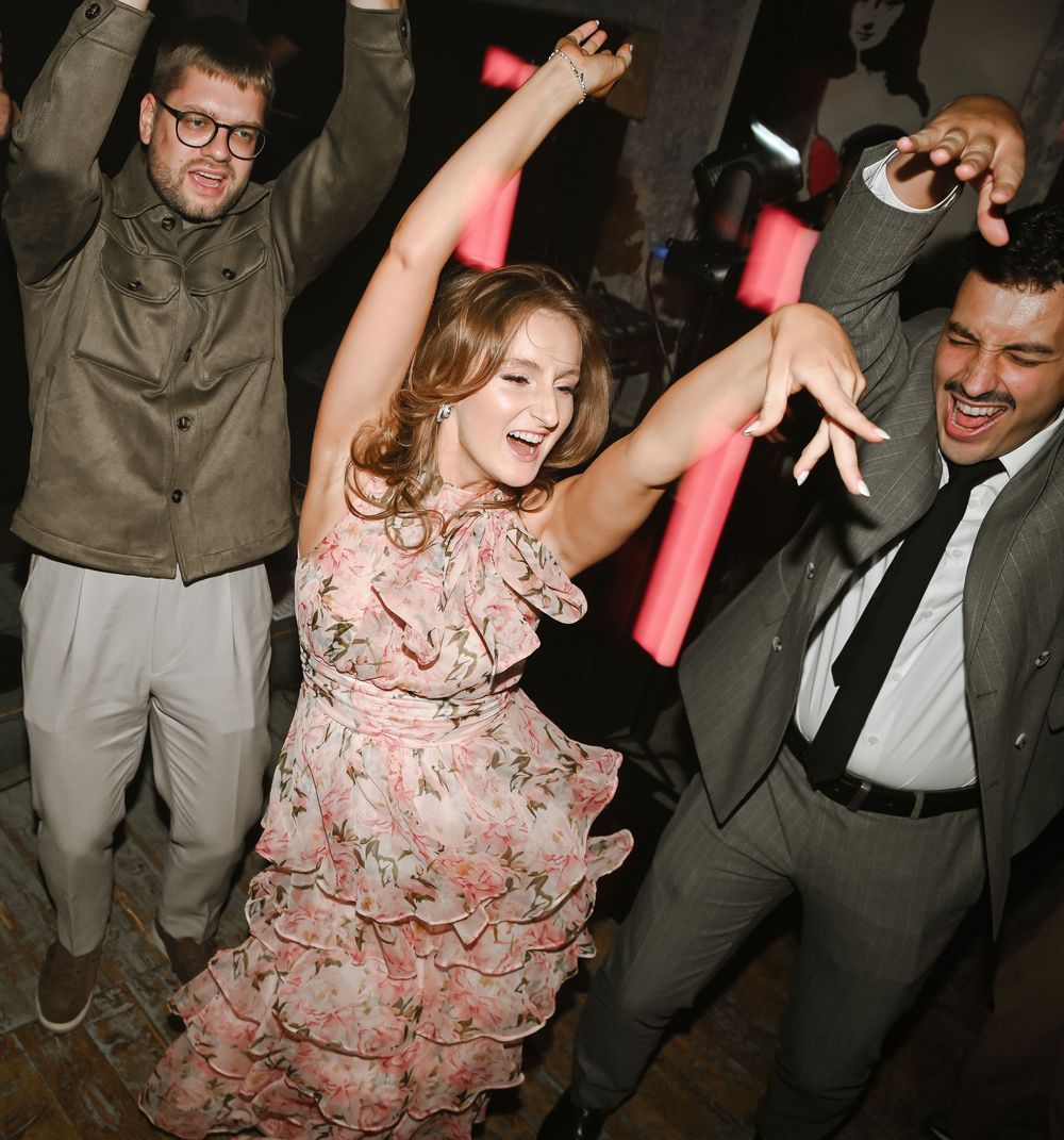 Three people dancing and celebrating at a party with someone wearing a pink floral dress in the center.