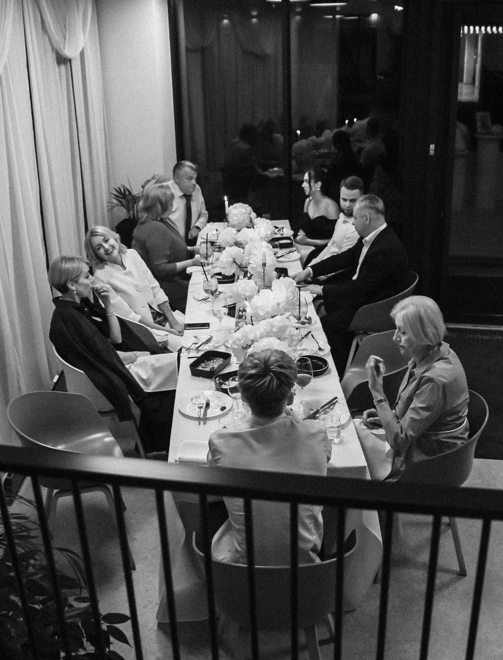 A black and white photograph of people dining together at a long table in an elegant restaurant setting.