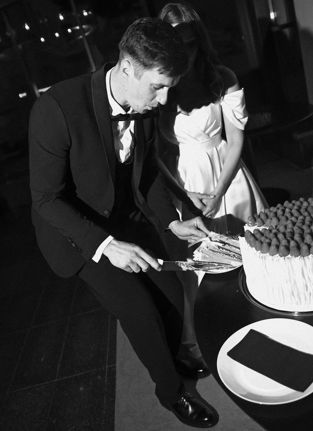 A man in a tuxedo signs something while standing next to a formal event table in black and white.