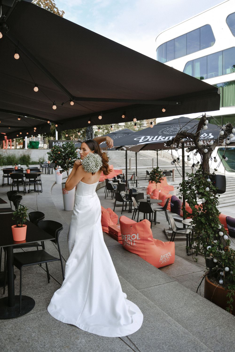 Elegant bride in white dress stands on outdoor rooftop terrace with string lights and modern city views.