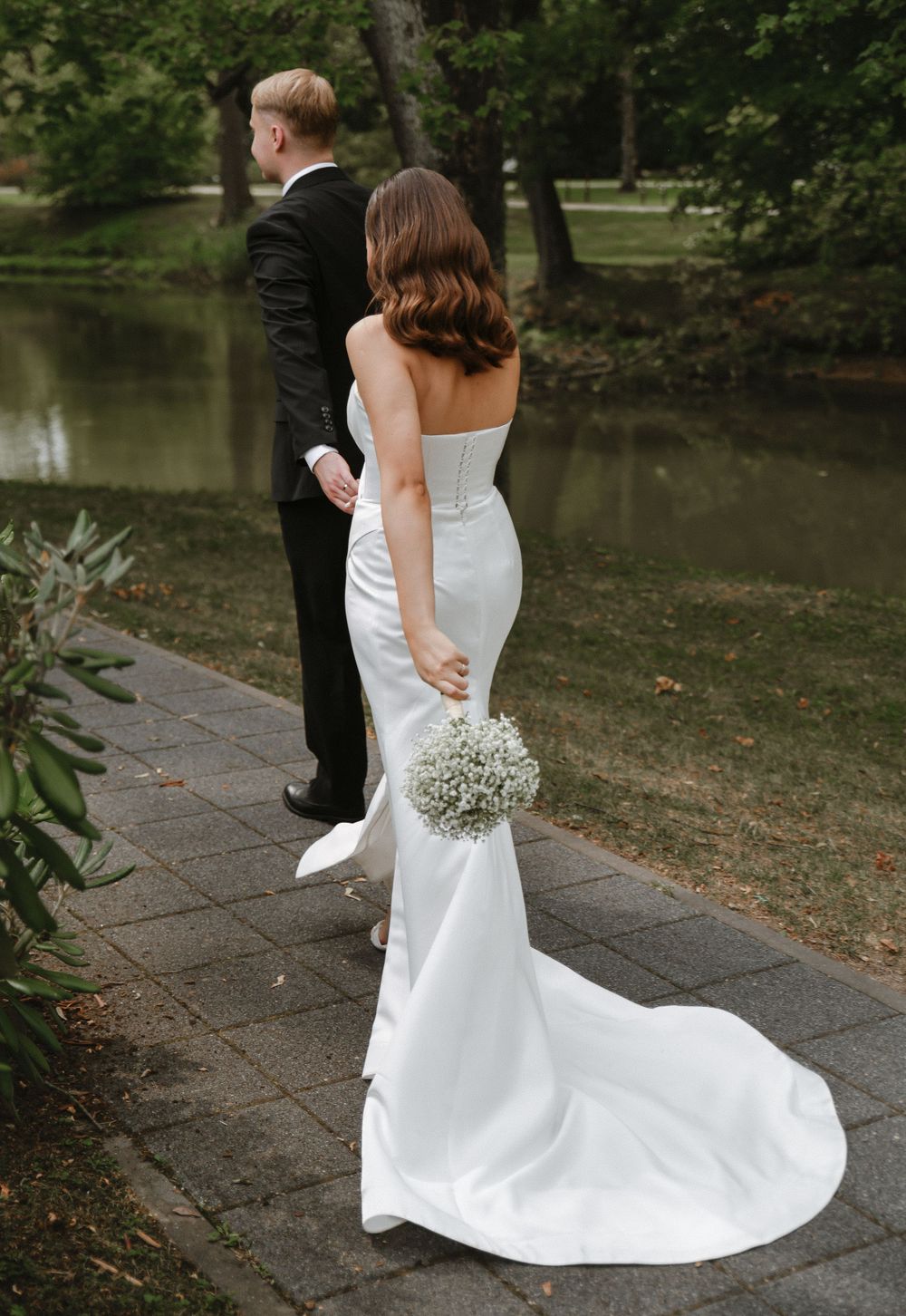 Elegant bride in flowing white gown walks along stone path near pond.