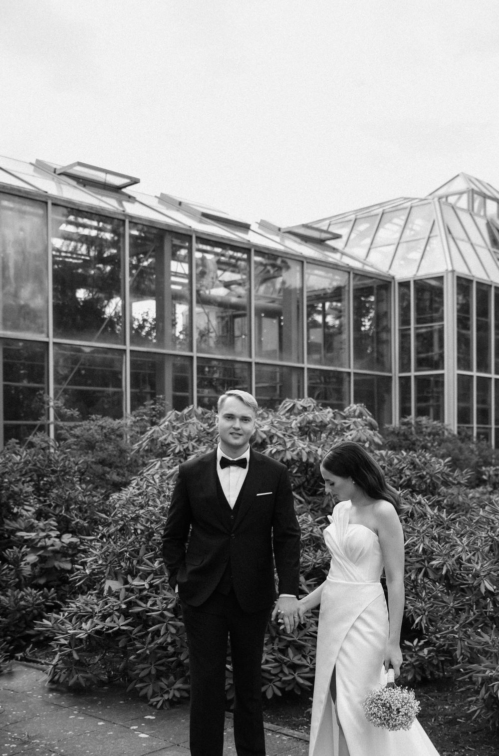 Wedding couple poses outside modern glass greenhouse building in black and white.