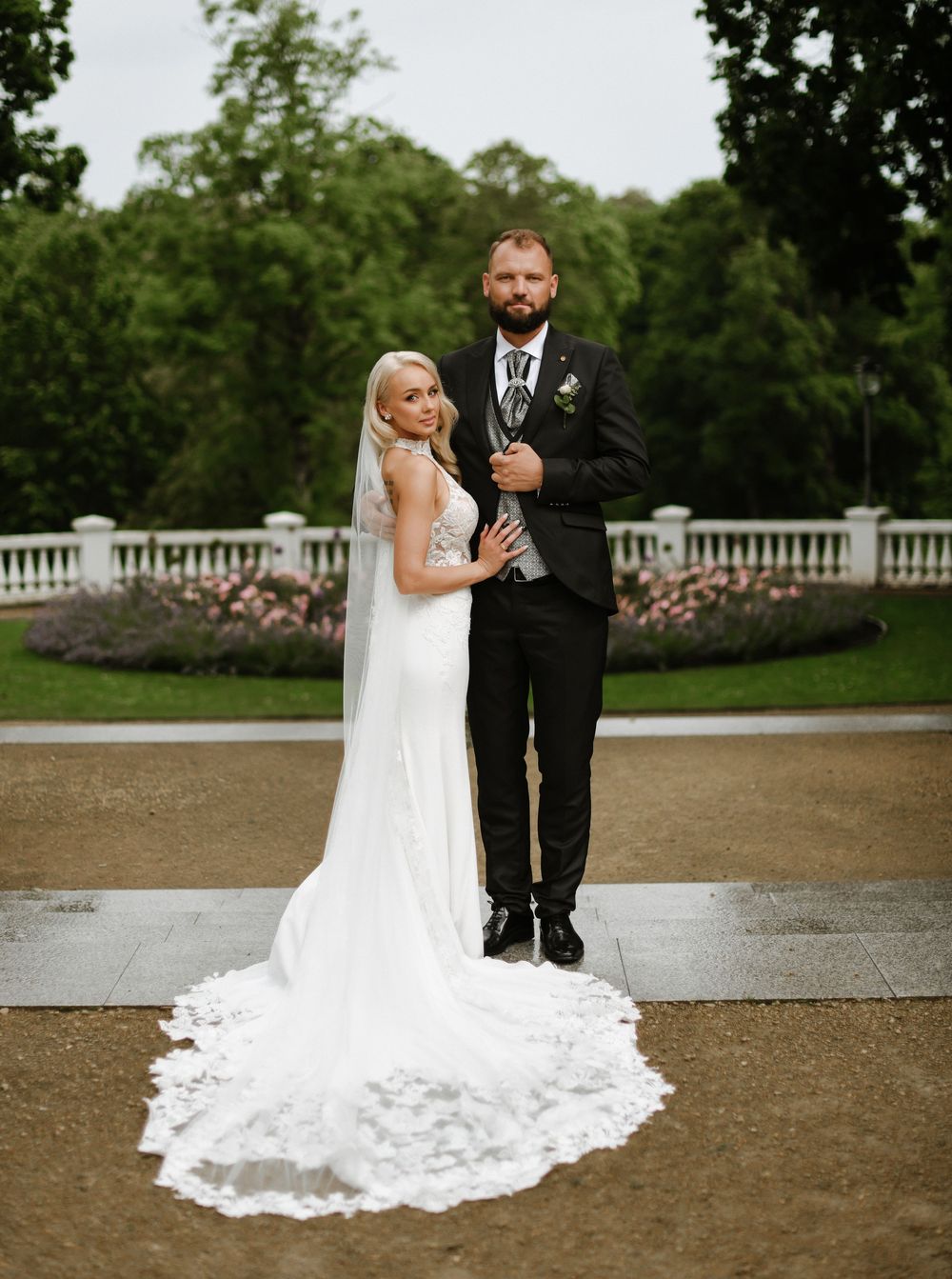 A wedding couple poses outdoors on a garden pathway with white railings and lush greenery in the background.
