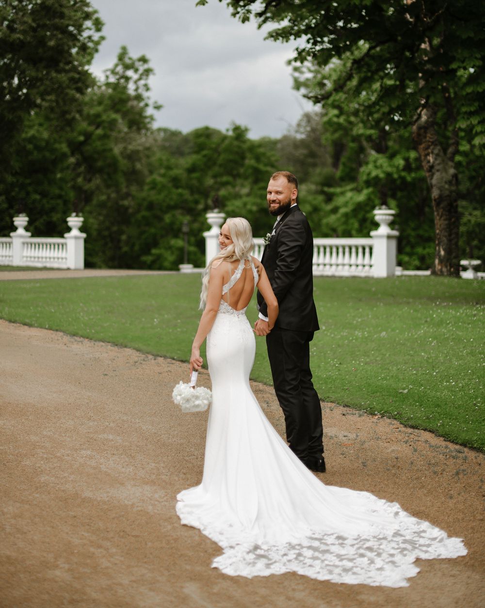 A bride and groom pose on a garden path with white fencing in the background, and beautiful white lace train on her dress.