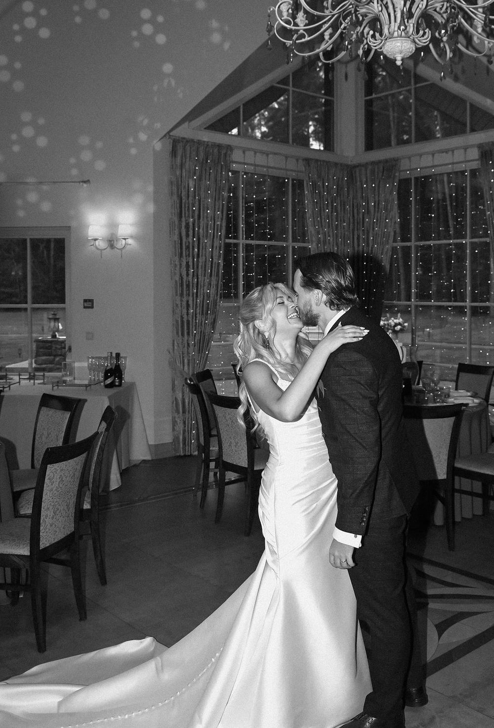 A couple dancing together in formal wedding attire inside an elegant reception venue with dim lighting and chandeliers.