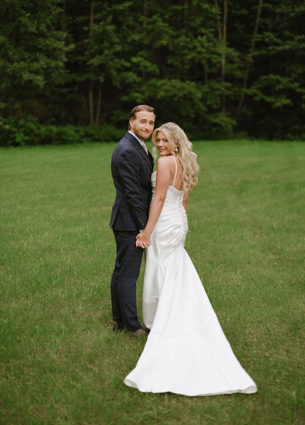 A couple in wedding attire dances in a romantic dip pose on a sunlit green lawn surrounded by trees.