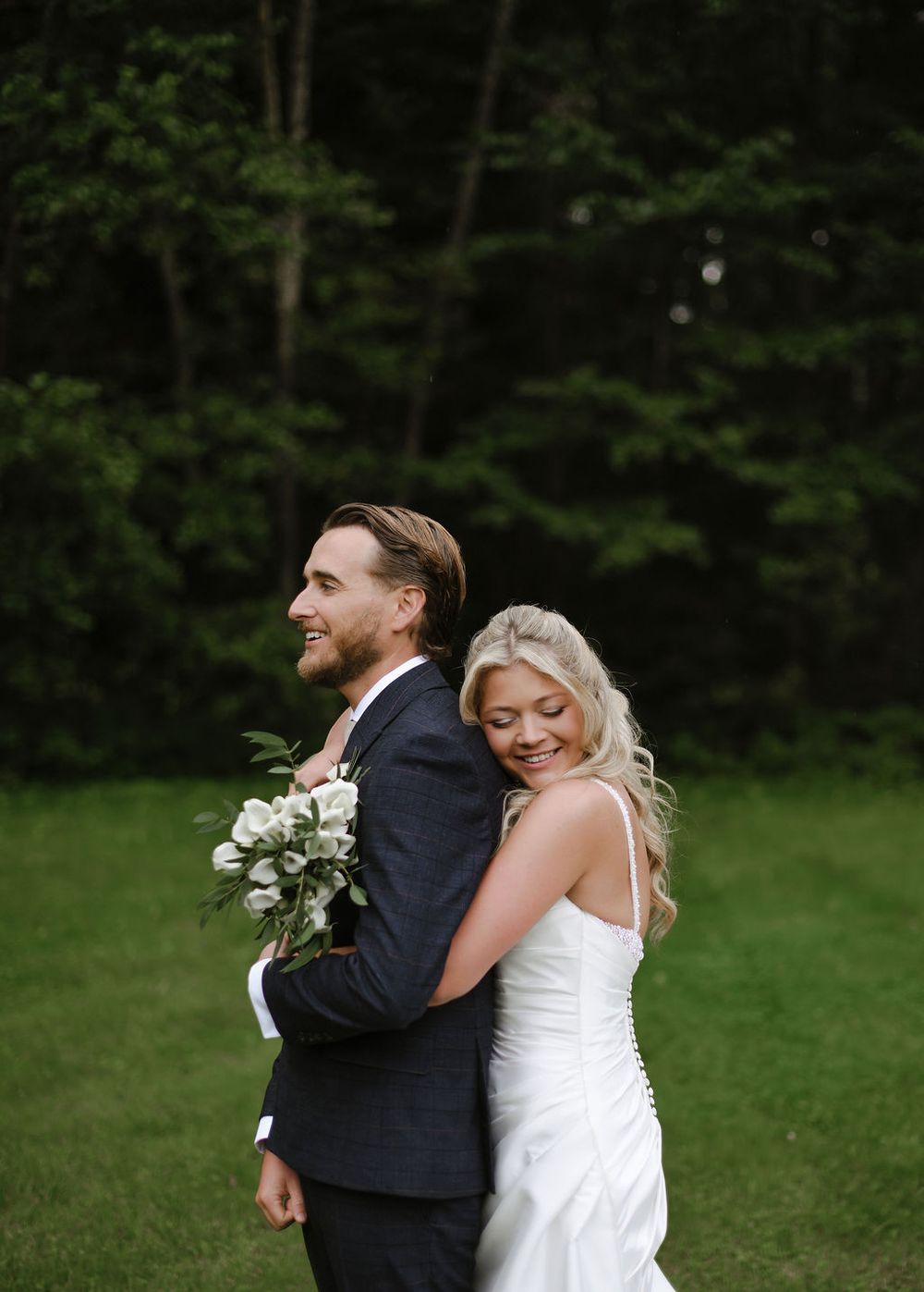 A joyful couple in wedding attire embraces and laughs together on a lush green lawn with forest backdrop.