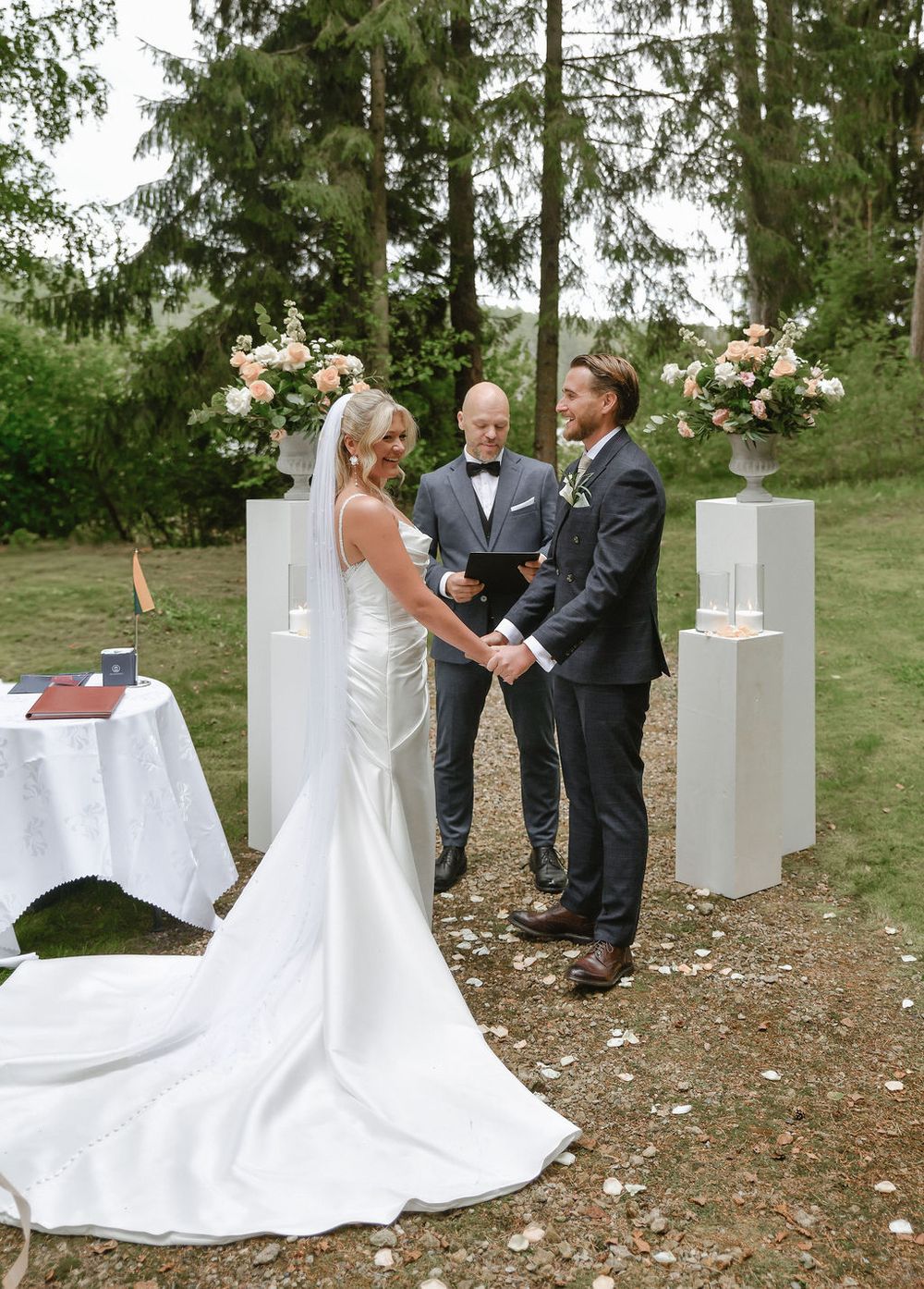 Wedding ceremony sequence outdoors with white decor elements and evergreen trees in the background.