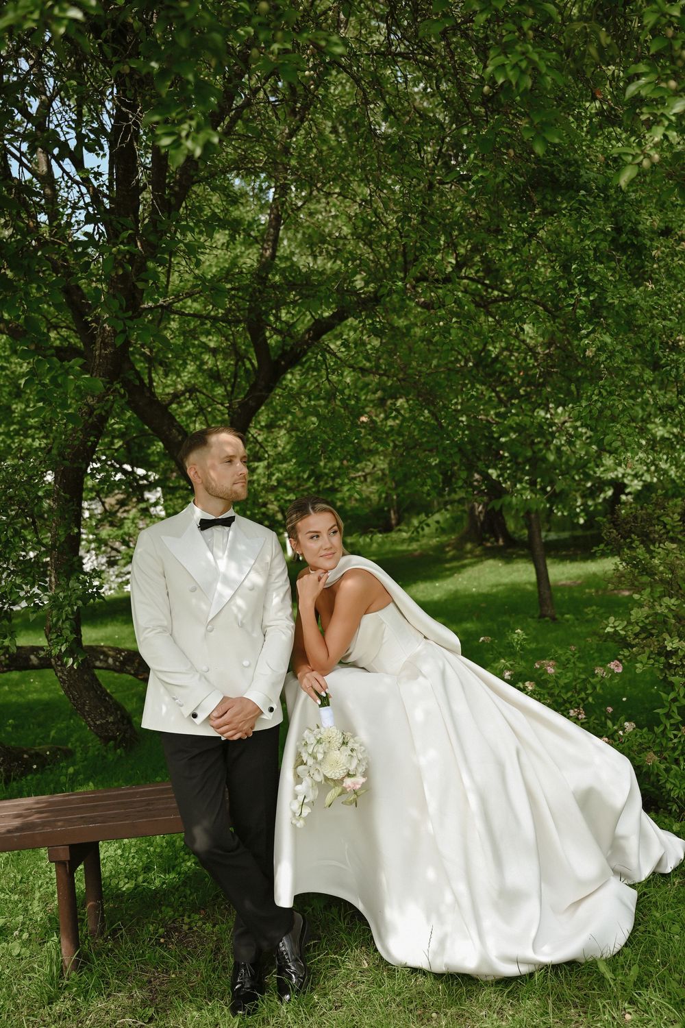 Bride and groom pose outdoors under lush green trees, bride in white gown holding bouquet, groom in white tuxedo.
