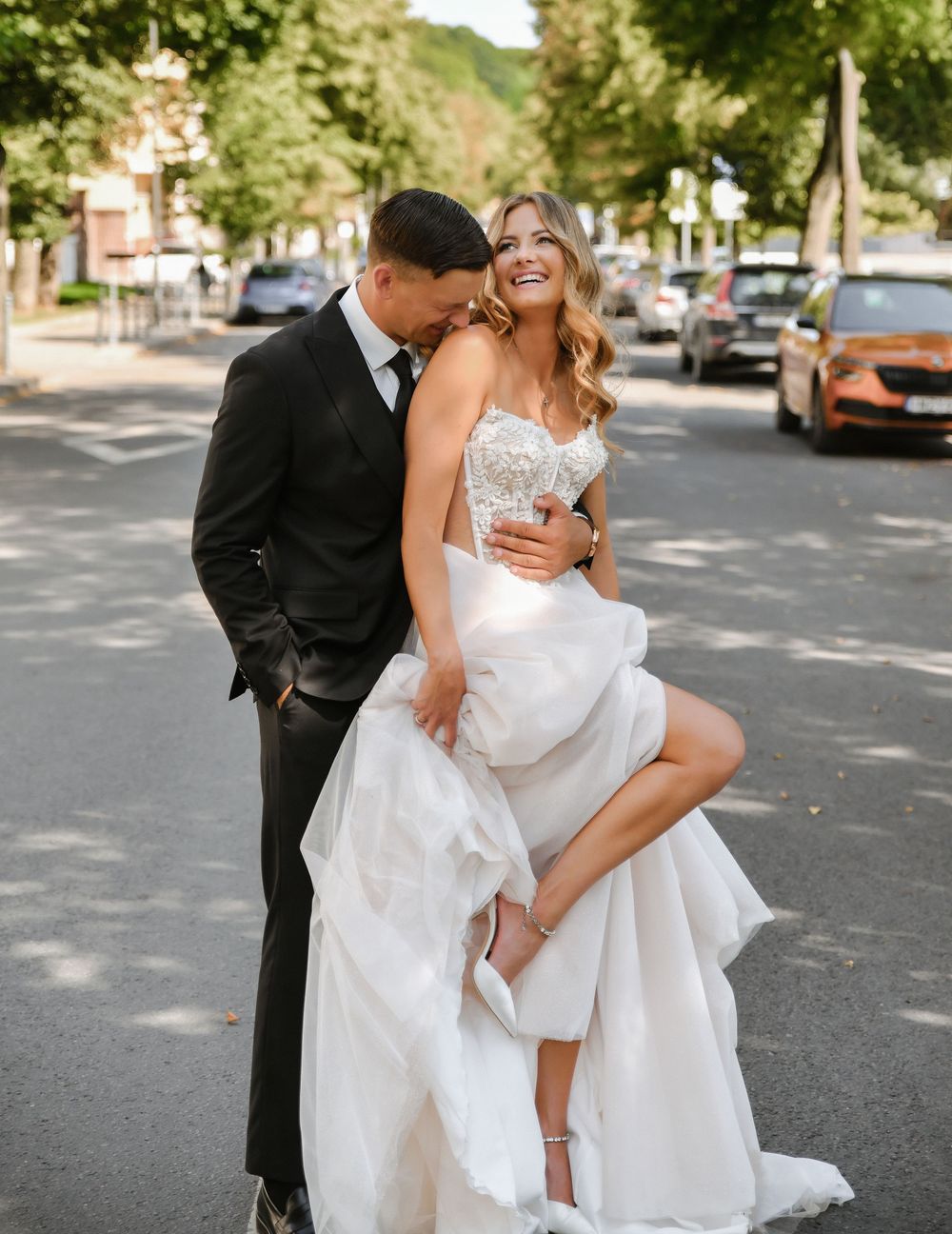 A couple in formal wedding attire share an intimate moment on a tree-lined street.