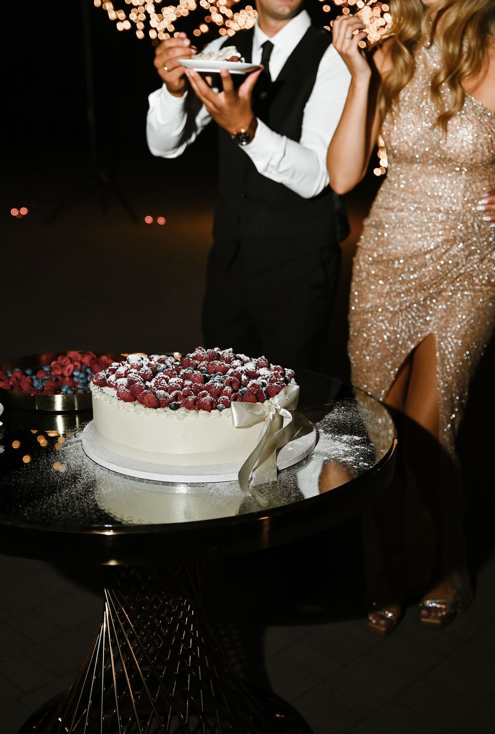 A elegant white cake decorated with red berries sits on a dark table while partygoers in formal attire celebrate nearby.