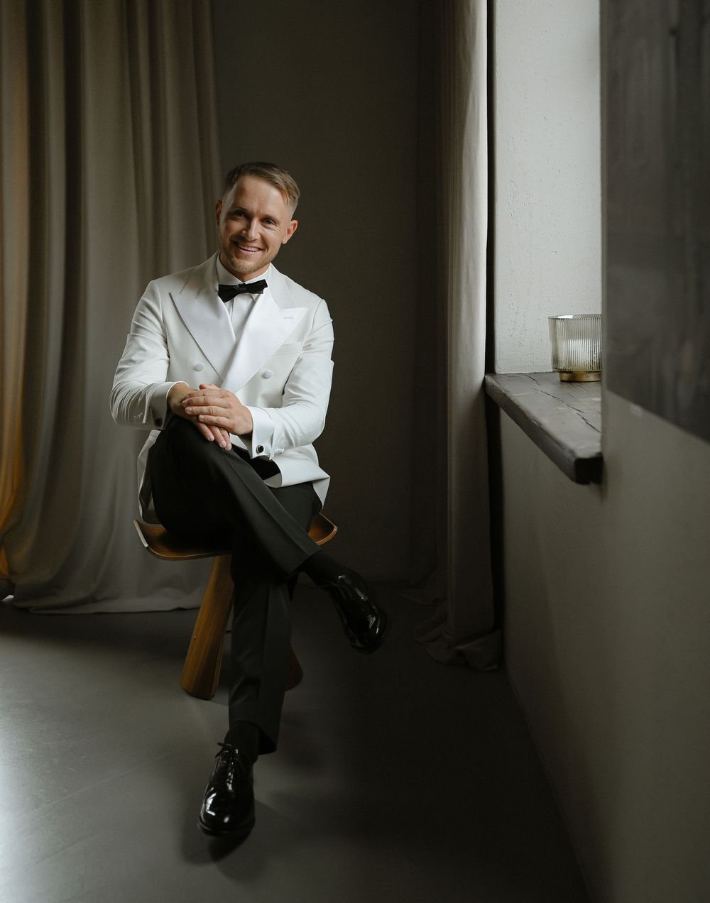 Smiling man in white tuxedo jacket and bow tie seated elegantly in a dimly lit room.
