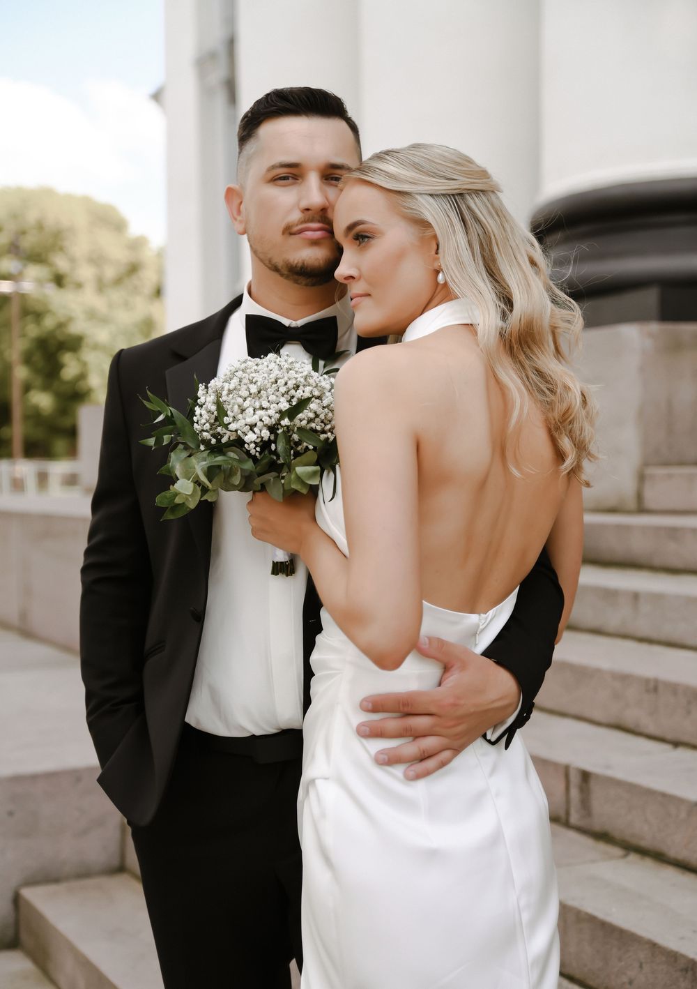 Newlywed couple embraces on stone steps, bride in white gown holds baby's breath bouquet while groom wears classic tuxedo.