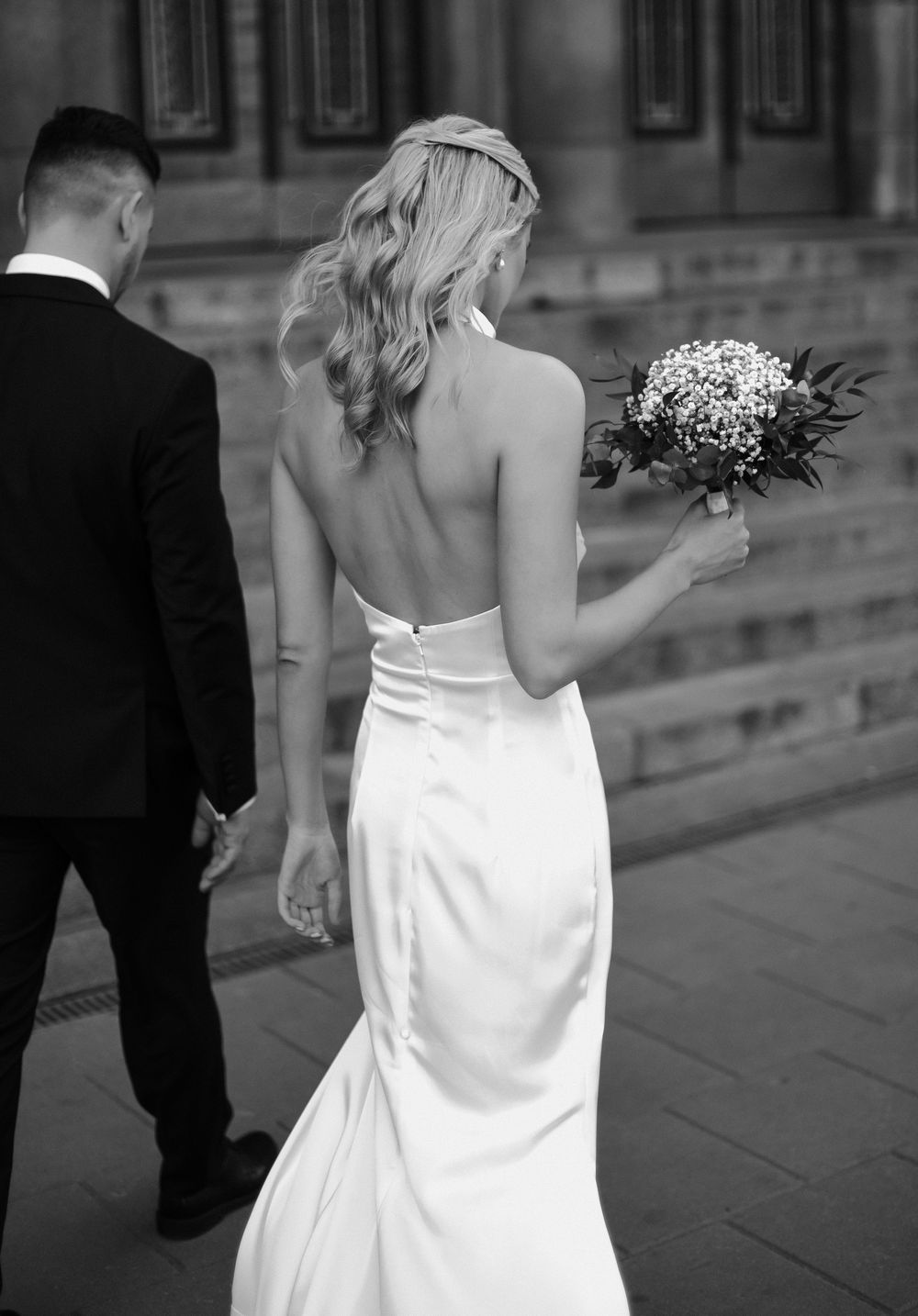 Black and white wedding photo showing bride in backless white dress holding bouquet walking alongside groom in dark suit.
