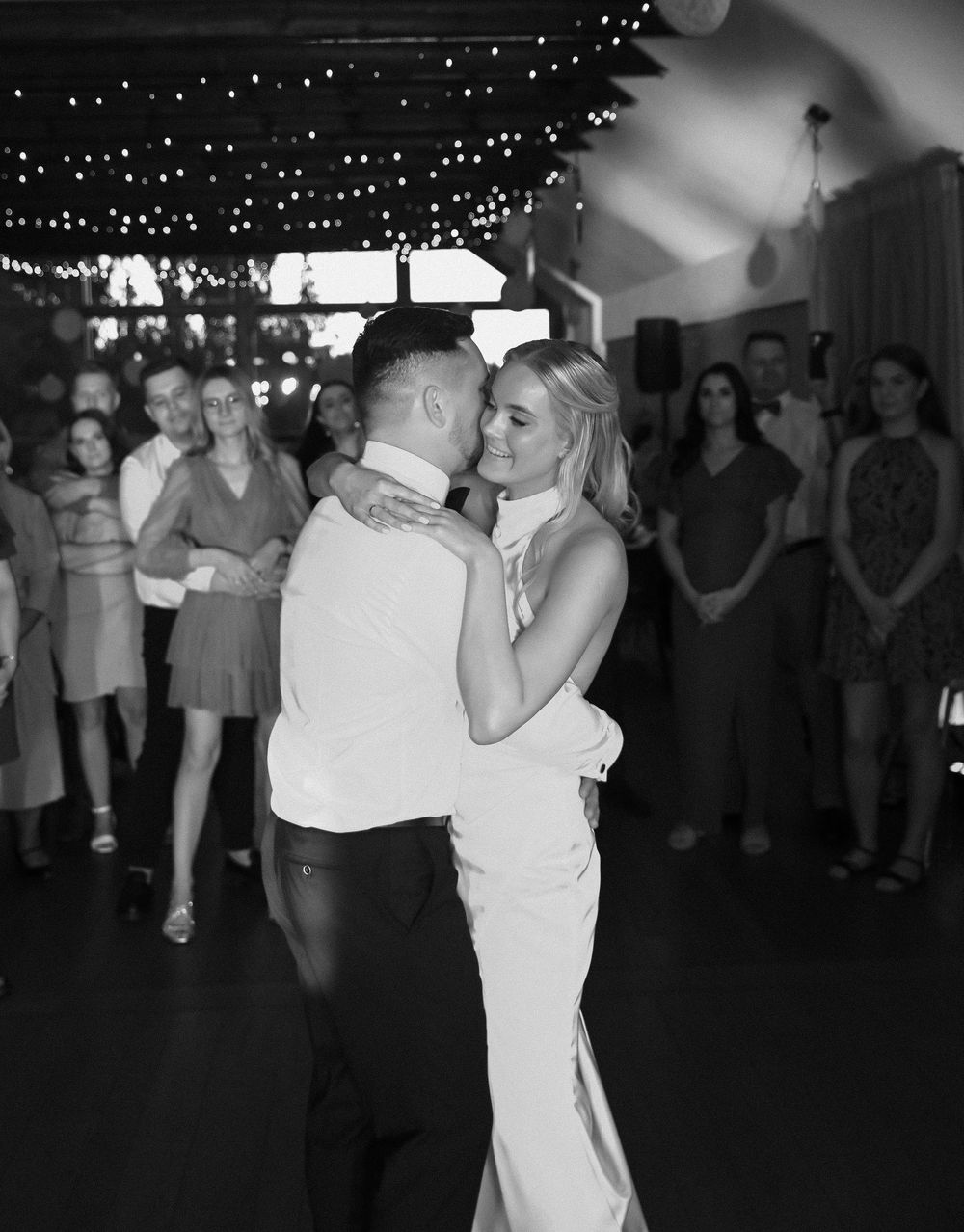 A romantic first dance at a wedding reception under twinkling string lights while guests watch in the background.