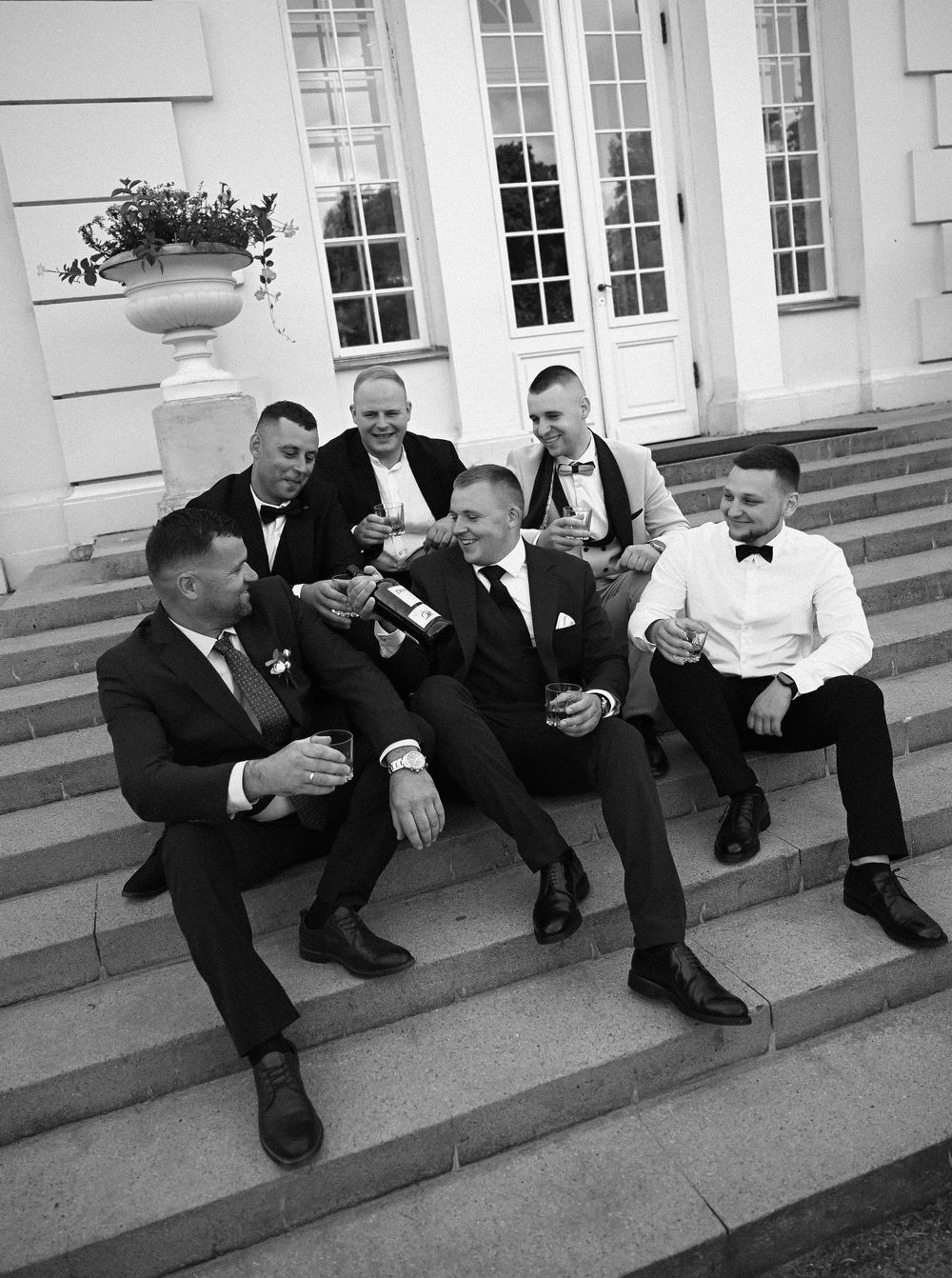 Groomsmen in black tuxedos sitting on elegant venue steps, laughing and celebrating together in a classic black and white photo.