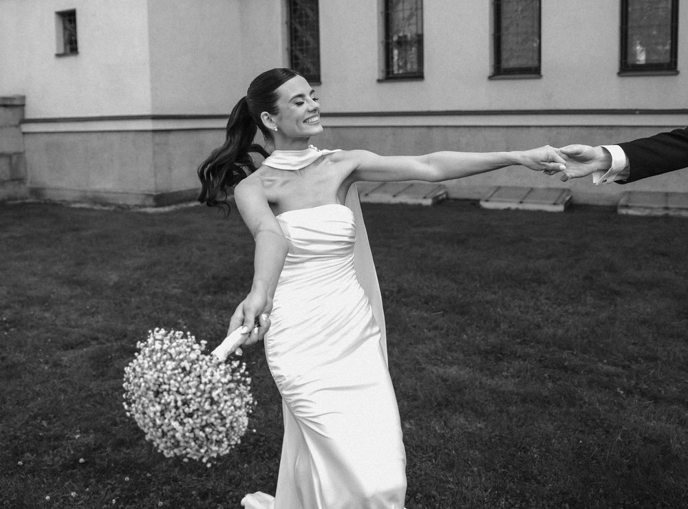 Elegant bride in white dress swirls and dances gracefully on outdoor lawn near white building.