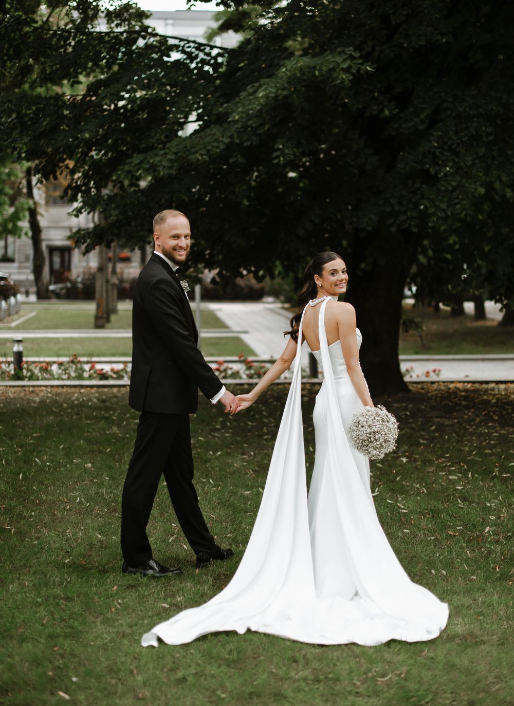 A couple in wedding attire holds hands while walking on grass with lush trees in the background.