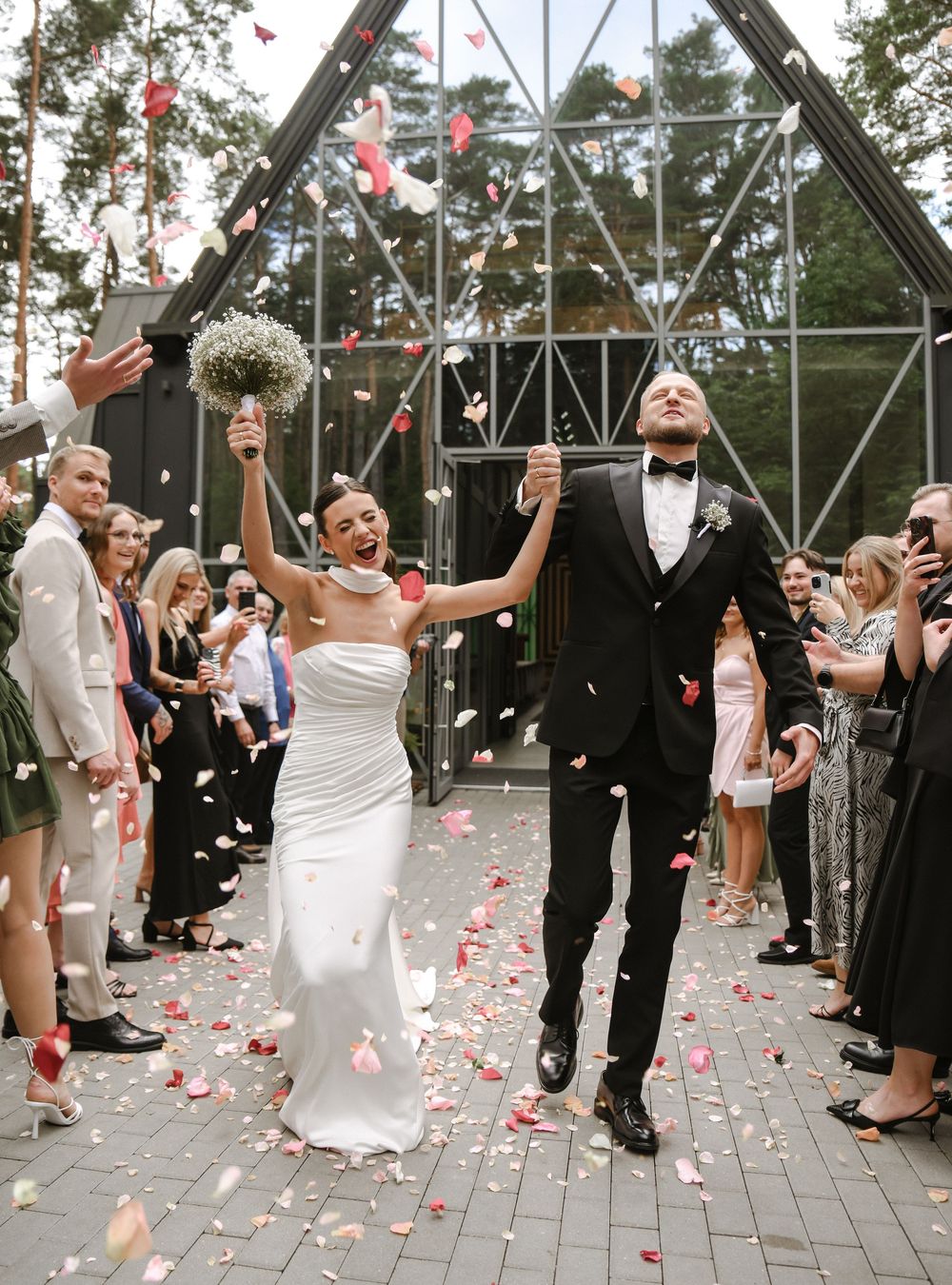 A couple celebrates their wedding ceremony under a glass greenhouse while guests shower them with pink rose petals.