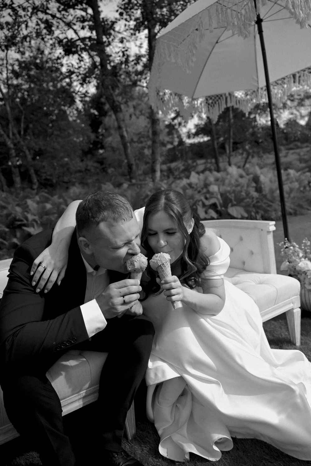 Black and white photo of bride and groom sharing ice cream cones on a white sofa outdoors at their wedding reception.